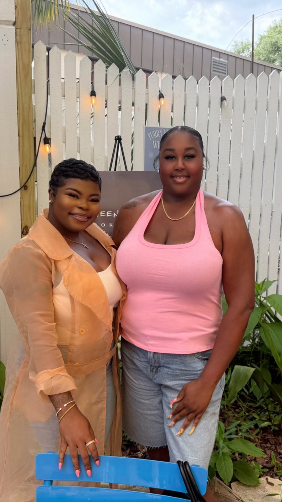 Two women smiling in front of a white picket fence