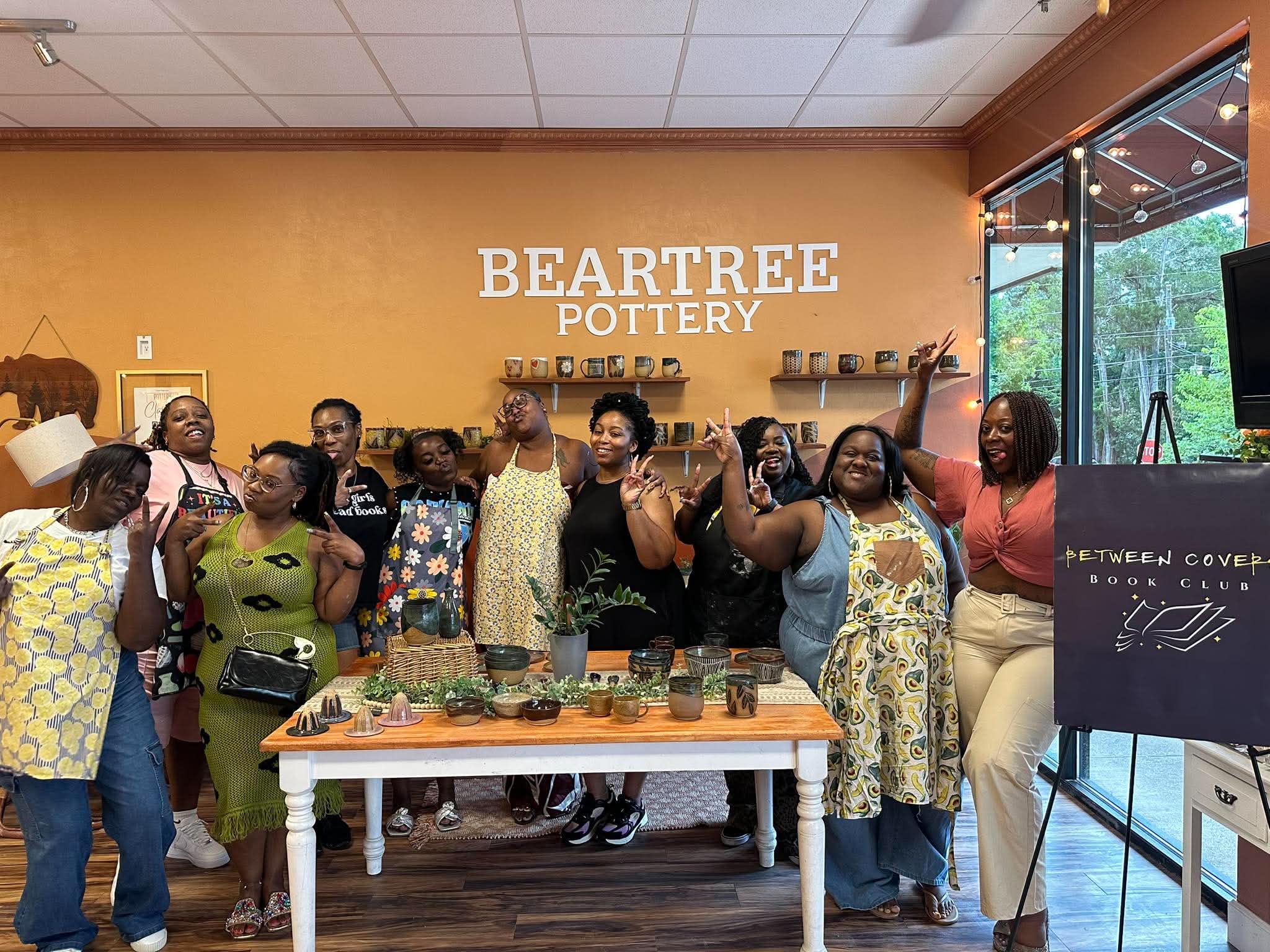 Between Covers members posing around a table of handmade pottery at Beartree Pottery with author Keyy Davis, next to the club's sign